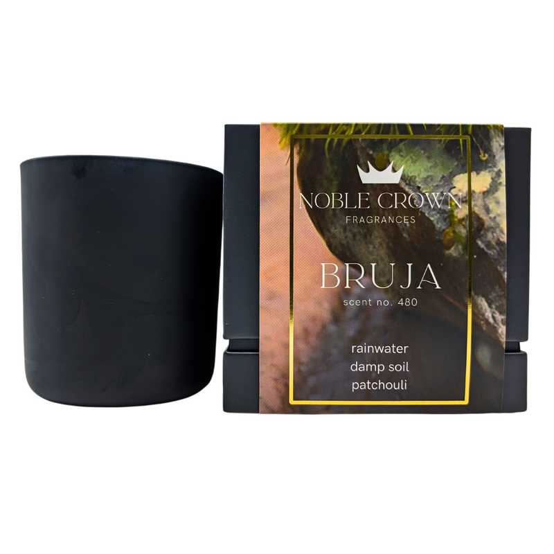 A black candle with a label that reads 'BRUJA' and indicates the scent is a witchy blend of rainwater, soil, and patchouli. The packaging has a crown and rainbow design on top.