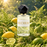 A clear glass perfume bottle labeled DUCHESS with a black faceted cap, surrounded by lemons and limes in a grassy outdoor setting with warm sunlight.