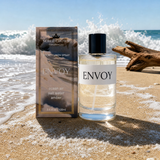 A bottle of Noble Crown Envoy Room spray and linen spray, along with its matching outer box featuring an ocean-themed design, are placed on a sandy beach, with waves and driftwood in the background.