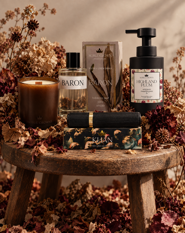A styled product arrangement on a rustic wooden stool surrounded by dried flowers and leaves. Visible items include a candle, a clear perfume bottle labeled BARON, a room spray labeled Baron, a soap bottle labeled HIGHLAND PLUM, and a perfume box labeled KHAN.