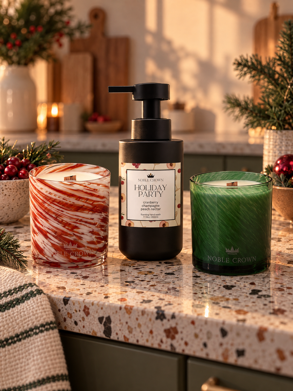 Three holiday-themed scented products from Noble Crown displayed on a speckled kitchen countertop: a red-and-white marbled candle, a black pump bottle labeled 'Holiday Party,' and a green candle. Festive greenery and candles are softly blurred in the background.