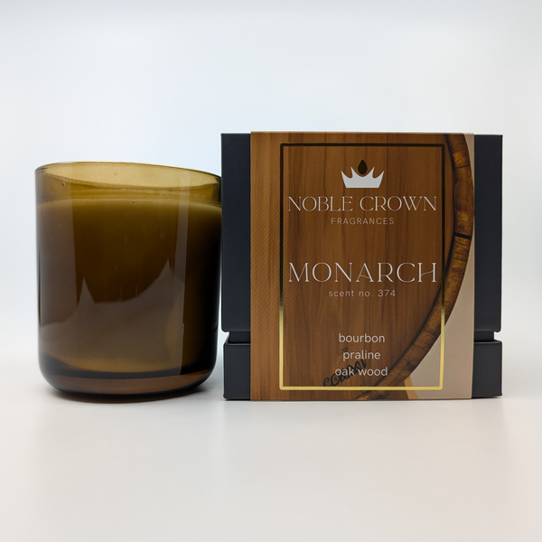 A brown candle next to its packaging box with the label Monarch and a scent description of bourbon, praline, amaretto, and oak wood.