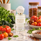 A clear glass perfume bottle labeled 'PICO DE GALLO' with a white faceted cap, styled on a marble surface with tomatoes, cilantro, peppers, and spice jars around it.