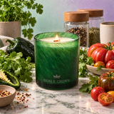 A green glass candle with a wick and visible flame sits on a marble surface surrounded by fresh herbs, tomatoes, peppers, and spice jars. The candle is branded NOBLE CROWN.