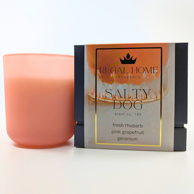 A pink candle next to its packaging, which has 'SALTY DOG' written on it, with additional text indicating the fragrance components such as fresh rhubarb, pink grapefruit, and geranium.